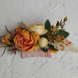 Op de afbeelding: Een bloemenhaar kam met oranje, gele en witte bloemen. De kam is gemaakt van koperdraad en heeft een delicaat ontwerp.