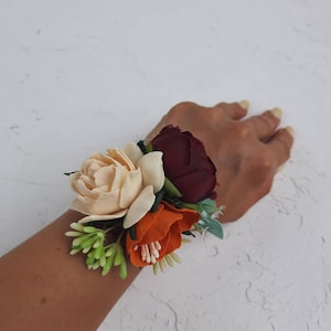May include: A wrist corsage with three silk flowers. The flowers are a cream rose, a burgundy rose, and an orange flower. The flowers are surrounded by green leaves.