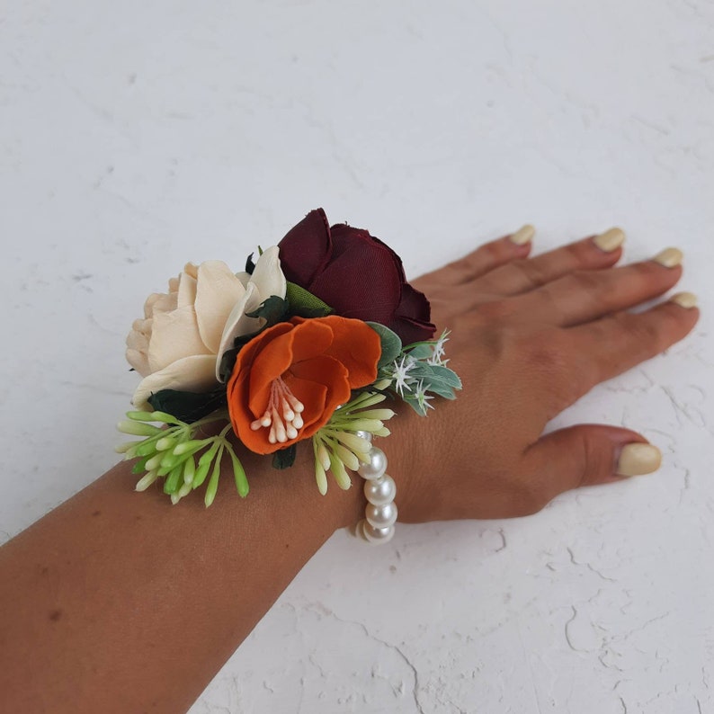 May include: A wrist corsage with a burgundy, cream, and orange flower arrangement. The flowers are accented with greenery and a pearl bracelet.