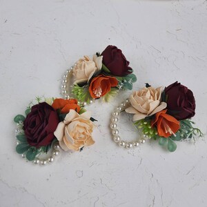 May include: Three floral wrist corsages with burgundy, peach, and cream colored fabric flowers. Each corsage is accented with pearl beads and greenery.