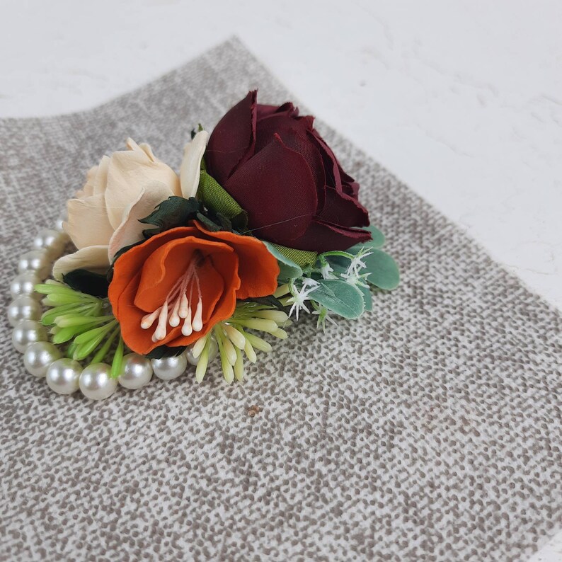 May include: A floral arrangement with three artificial flowers. The flowers are a burgundy rose, a cream-colored rose, and an orange flower. The flowers are arranged on a pearl bracelet.