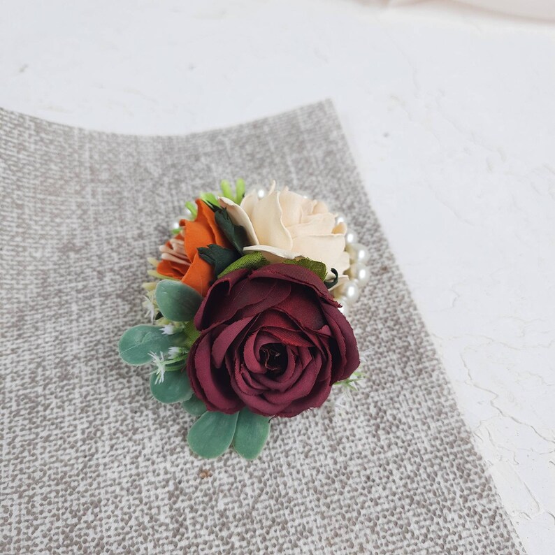 May include: A burgundy, cream, and orange fabric flower boutonniere with pearl accents.
