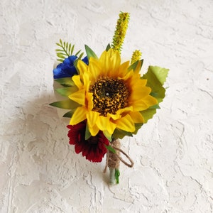 May include: A small bouquet of artificial flowers with a yellow sunflower, red and blue flowers, and green leaves. The flowers are tied together with twine.