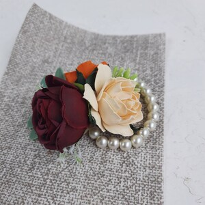May include: A burgundy rose, a cream rose, and an orange rose are arranged together with a pearl necklace. The flowers are on a gray textured background.