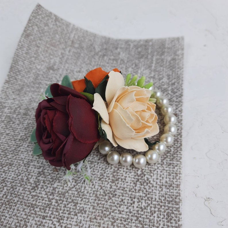 May include: A burgundy rose, a cream rose, and an orange rose are arranged together with a pearl necklace. The flowers are on a gray textured background.