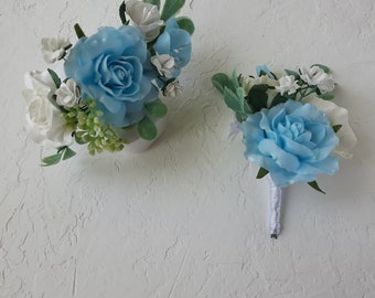 Puede incluir: Un arreglo floral con una rosa grande de color azul claro, rosas blancas más pequeñas, hojas verdes y pequeñas flores blancas, junto a un boutonniere a juego con una rosa azul claro y un tallo envuelto en cinta blanca.