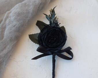 May include: A black fabric flower boutonniere featuring layered petals, a central cluster of small black beads, dark green leaves, and a small sprig of greenery. The stem is wrapped in black ribbon, and a black satin bow is tied at the base of the flower.