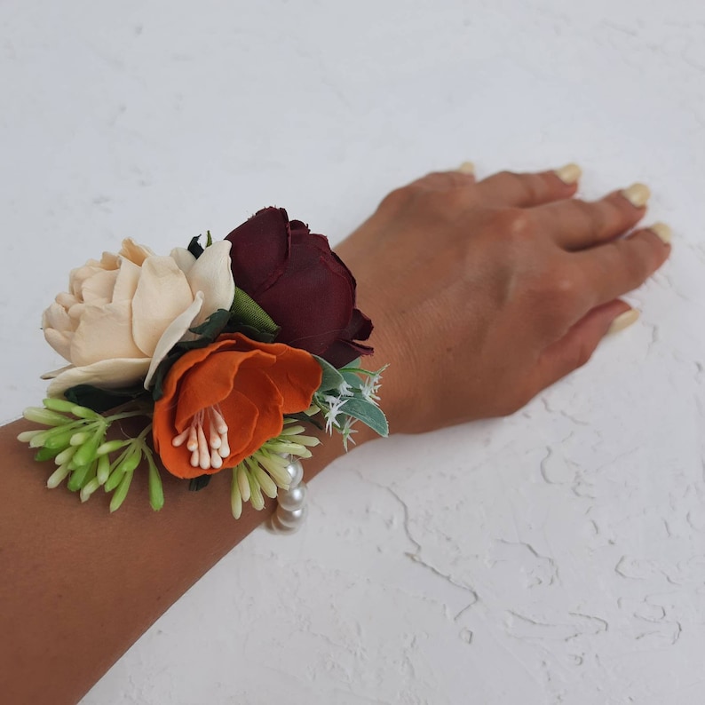 May include: A wrist corsage with a burgundy, cream, and orange flower arrangement. The flowers are accented with green leaves and a pearl bracelet.