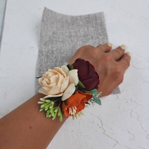 May include: A wrist corsage with three fabric flowers. The flowers are cream, orange, and burgundy. The corsage is accented with green leaves.