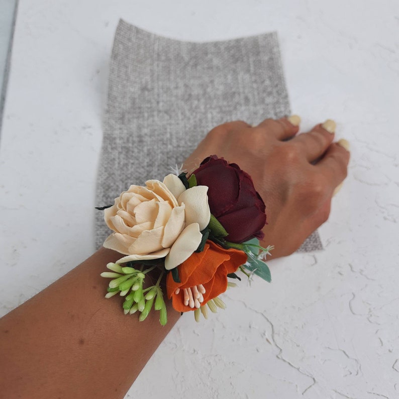 May include: A wrist corsage with three fabric flowers. The flowers are cream, orange, and burgundy. The corsage is accented with green leaves.