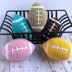 May include: Five crocheted footballs in a black wire basket. The footballs are in different colors: brown, yellow, pink, teal, and beige.