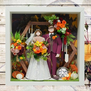 May include: A papercraft wedding scene with a bride and groom in a white frame. The bride is wearing a white dress with lace details and holding a bouquet of orange, yellow, and red flowers. The groom is wearing a burgundy suit and a white shirt with a vest. The scene is set in a forest with a wooden archway and greenery.