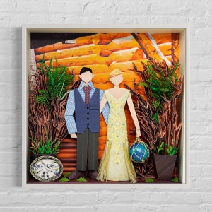 Può includere: Un diorama di cartapesta che rappresenta una coppia di sposi in piedi davanti a una baita di tronchi. La sposa indossa un abito giallo e lo sposo indossa una camicia a quadri blu e un gilet. Il diorama è incorniciato in bianco.