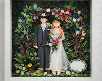 Puede incluir: Una obra de arte en papel 3D enmarcada que muestra a una pareja sin rostro bajo un arco floral. La figura de la izquierda viste un traje gris oscuro y la de la derecha un vestido blanco con un ramo. El texto KYARTSTUDIO aparece en la base.