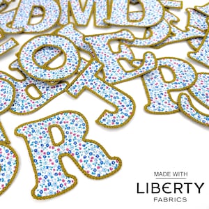 Könnte beinhalten: Ein Stapel Stoffbuchstaben in einem blauen und weißen Blumenmuster mit goldener Umrandung. Die Buchstaben sind in zufälliger Reihenfolge angeordnet und alle in Großbuchstaben. Die Buchstaben sind aus Liberty-Stoffen gefertigt und perfekt für Bastelprojekte.