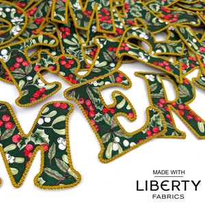 May include: A close-up of a pile of fabric letters spelling out "Merry" in a green and white floral pattern with red berries. The letters are outlined in gold thread and have a vintage feel. The text "MADE WITH LIBERTY FABRICS" is visible at the bottom of the image.