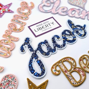 Könnte beinhalten: Eine Auswahl an Namensaufnähern und Sternformen aus Stoff mit verschiedenen Blumen- und Sternmustern. Die Namen sind in Schreibschrift gehalten und haben einen gestickten Rand. Ein Etikett lautet "MADE WITH LIBERTY FABRICS."