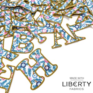 May include: A pile of fabric letters in a blue and pink floral pattern with gold trim. The letters are scattered on a white background. The letters spell out the word "LIBERTY" in gold lettering. The text "MADE WITH LIBERTY FABRICS" is visible in the bottom right corner of the image.