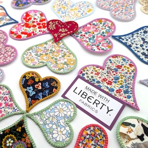 May include: A collection of handmade heart-shaped fabric patches featuring various floral patterns. The patches are made with Liberty fabrics and have a crocheted edge. The text "MADE WITH LIBERTY FABRICS." is displayed on a purple tag.