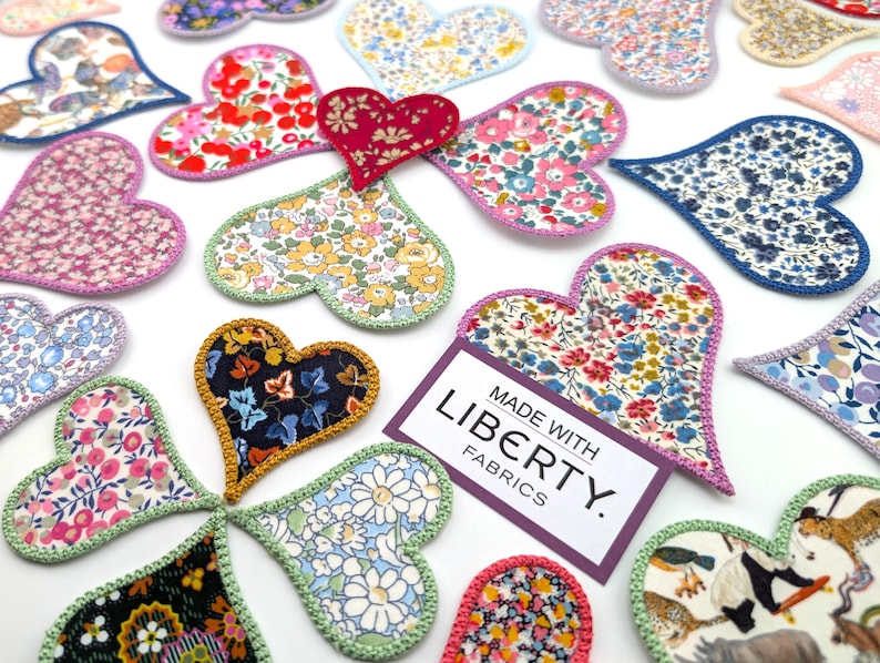 May include: A collection of handmade heart-shaped fabric patches featuring various floral patterns. The patches are made with Liberty fabrics and have a crocheted edge. The text "MADE WITH LIBERTY FABRICS." is displayed on a purple tag.