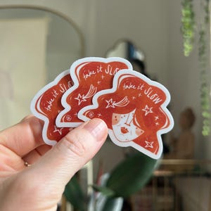 May include: Three red stickers with a white border are held in a hand. Each sticker features a stylized illustration of a woman's face with red hair and the text "take it SLOW" in white. Stars and crosses are also included in the design.