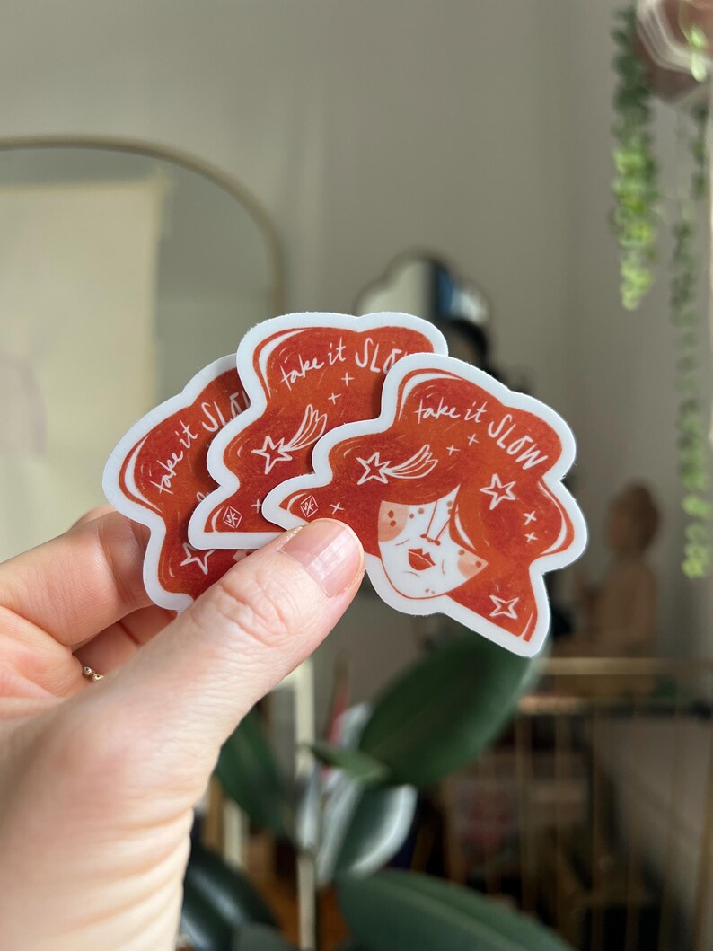 May include: Three red stickers with a white border are held in a hand. Each sticker features a stylized illustration of a woman's face with red hair and the text "take it SLOW" in white. Stars and crosses are also included in the design.