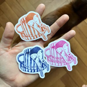May include: Three stickers featuring illustrations of a person's legs and torso sticking out of a coffee cup. The stickers are in orange, blue, and pink, with text that reads "More Espresso Less Depresso" and "kafe tanta".