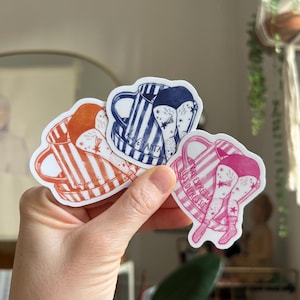 May include: Three coffee-themed stickers are held in a hand. The stickers feature illustrations of coffee mugs with legs. One is orange and white striped, one is blue and white striped, and one is pink. Text is included on the stickers.