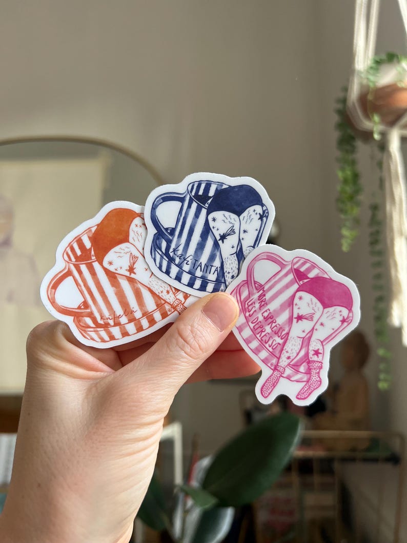 May include: Three coffee-themed stickers are held in a hand. The stickers feature illustrations of coffee mugs with legs. One is orange and white striped, one is blue and white striped, and one is pink. Text is included on the stickers.
