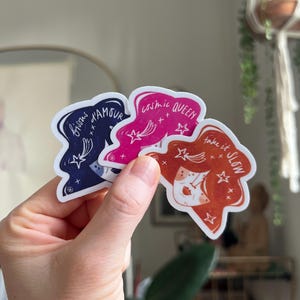 May include: Three die-cut stickers held in a hand. The stickers feature illustrations of women's faces with text: "bisous d'amour", "cosmic QUEEN", and "take it SLOW". The stickers are in shades of navy, pink, and orange.
