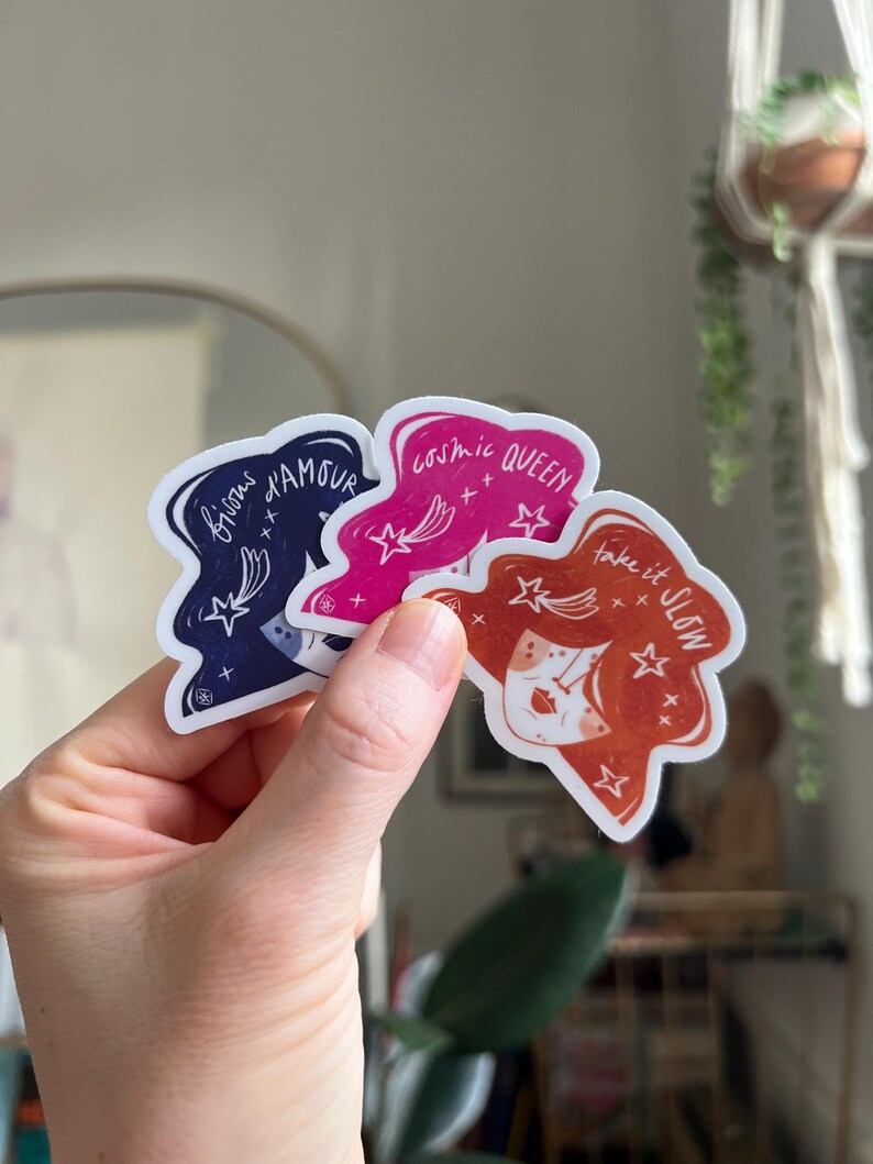 May include: Three die-cut stickers held in a hand. The stickers feature illustrations of women's faces with text: "bisous d'amour", "cosmic QUEEN", and "take it SLOW". The stickers are in shades of navy, pink, and orange.