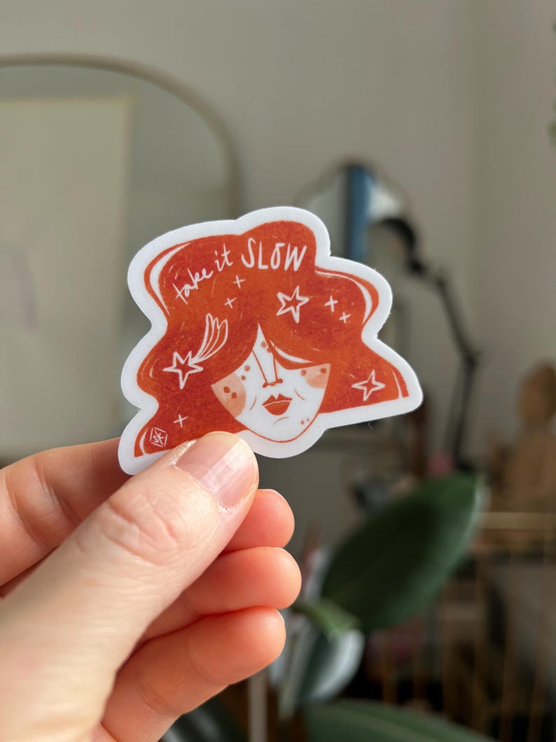 May include: A sticker featuring a stylized illustration of a woman's face with red hair and the words "take it SLOW". The design includes stars and a small logo. The sticker is held in a hand.