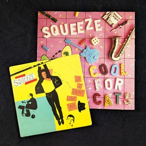 Könnte beinhalten: Zwei quadratische Musikalbum-Cover. Das hintere Cover ist rosa mit einem Gittermuster und zeigt den Text SQUEEZE COOL FOR CATS, eine blaue Miniaturgitarre, ein goldenes Saxophon, einen Würfel und kleine Pillen. Das vordere Cover ist gelb.
