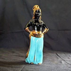 May include: A ceramic figurine of a standing figure with a black upper body, gold accents on the head, waist, and arms, and a long, flowing turquoise skirt. The figure has hands placed on the hips and a smooth, glossy finish throughout the piece.