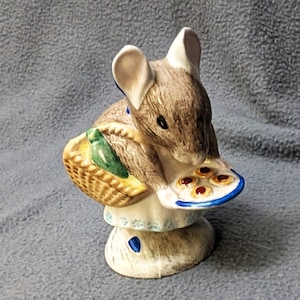 May include: A ceramic figurine of a brown mouse wearing a white apron with blue floral details. The mouse carries a woven basket on its back and holds a white plate with four small round cookies featuring red centers in its front paws.