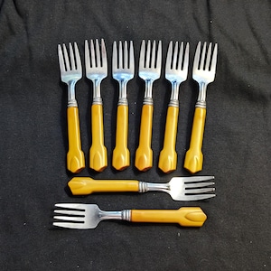 May include: A set of eight stainless steel forks featuring mustard yellow plastic handles. Six forks are arranged vertically in a row, while two additional forks are positioned horizontally below them. Each handle has a distinct flared end.