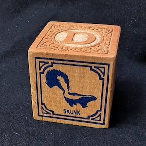May include: A wooden alphabet block featuring the letter D on the top surface within a decorative border. The front face displays a dark blue graphic of a skunk with the word SKUNK printed in dark blue capital letters below the animal.