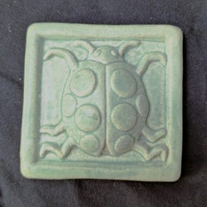 May include: A square, light green ceramic tile featuring a raised, embossed design of a ladybug in the center. The ladybug has a rounded body with visible circular spots and six legs extending outward, all finished in a matte, monochromatic glaze.