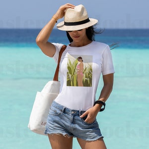 Puede incluir: Camiseta blanca con un gr&aacute;fico de una mujer con un traje de ba&ntilde;o rosa de pie frente a plantas verdes altas. La persona lleva un sombrero de paja y gafas de sol. La persona tambi&eacute;n lleva pantalones cortos de mezclilla y lleva un bolso de mano blanco.