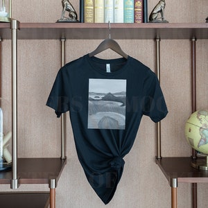 May include: Black t-shirt with a landscape painting on the front. The artwork is a grayscale depiction of a coastal scene. The shirt is hanging on a wooden hanger against a neutral background, showcasing a minimalist aesthetic.