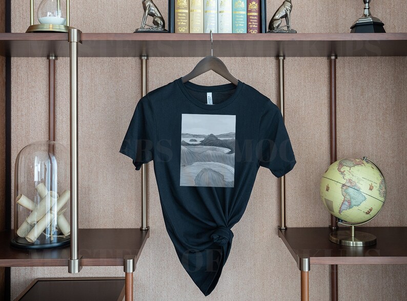 May include: Black t-shirt with a landscape painting on the front. The artwork is a grayscale depiction of a coastal scene. The shirt is hanging on a wooden hanger against a neutral background, showcasing a minimalist aesthetic.