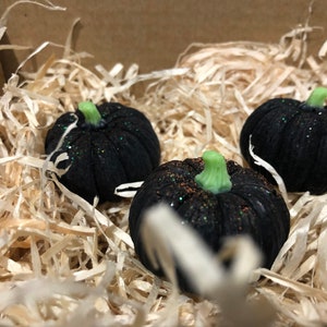 K&ouml;nnte beinhalten: Drei kleine schwarze K&uuml;rbisse mit gr&uuml;nen Stielen und Glitzerakzenten liegen in zerkleinertem Papier.