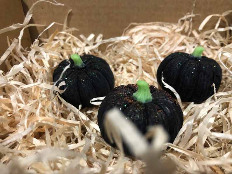 K&ouml;nnte beinhalten: Drei kleine schwarze K&uuml;rbisse mit gr&uuml;nen Stielen und Glitzerakzenten liegen in zerkleinertem Papier.