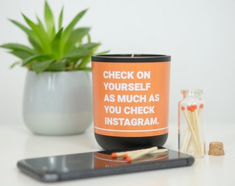Könnte beinhalten: Orangefarbene Kerze mit schwarzer Aufschrift "Check on yourself as much as you check Instagram." Die Kerze steht auf einer weißen Oberfläche mit einem kleinen Glas mit Streichhölzern und einem Korken.