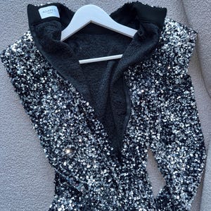May include: A black sequined jacket with a high collar and a black interior. The jacket is hanging on a white hanger. The brand name "AGAPES WEAR" is visible on the collar.