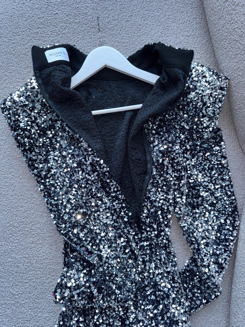 May include: A black sequined jacket with a high collar and a black interior. The jacket is hanging on a white hanger. The brand name "AGAPES WEAR" is visible on the collar.