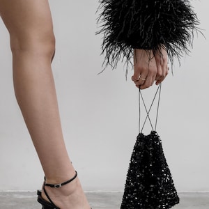 Può includere: Una borsa da sera nera con paillettes e un lungo cinturino nero. La borsa è tenuta da una persona che indossa tacchi neri con paillettes e un abito nero con bordi di piume.