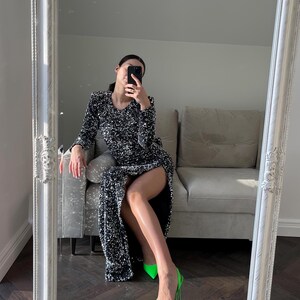 May include: A woman wearing a silver sequined dress with a thigh-high slit and neon green high heels. She is sitting on a light gray couch and holding a black smartphone.