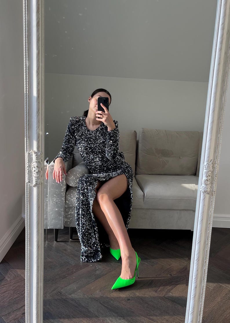 May include: A woman wearing a silver sequined dress with a thigh-high slit and neon green high heels. She is sitting on a light gray couch and holding a black smartphone.