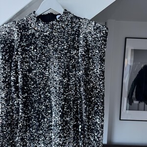 May include: A black dress covered in silver sequins hangs on a white hanger. The dress has long sleeves and a high neckline. The sequins create a shimmering effect, reflecting light. The dress is a formal garment.
